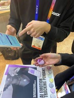 A PieX worker and visitor at the PieX booth at CES 2025 talking about the PieX AI Emotion Tracking Pendant. The visitor is holding the circular pendant, that's purple, in her hand while holding a brochure on the technology. A PieX worker and visitor at the PieX booth at CES 2025 talking about the PieX AI Emotion Tracking Pendant. The visitor is holding the circular pendant, that's purple, in her hand while holding a brochure on the technology.