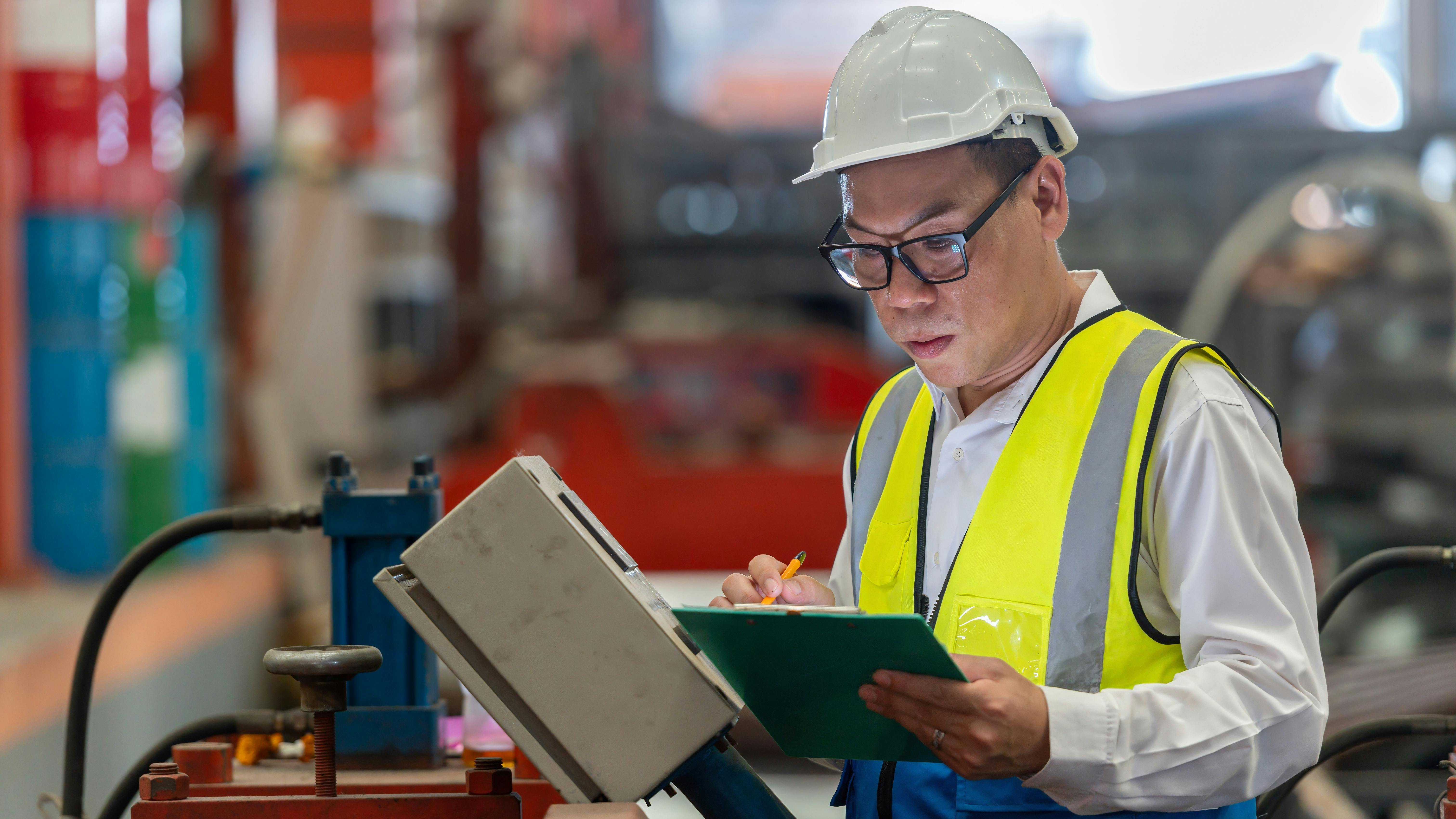Asian male engineer in uniform inspecting warehouse and checking heavy machinery in industrial factory