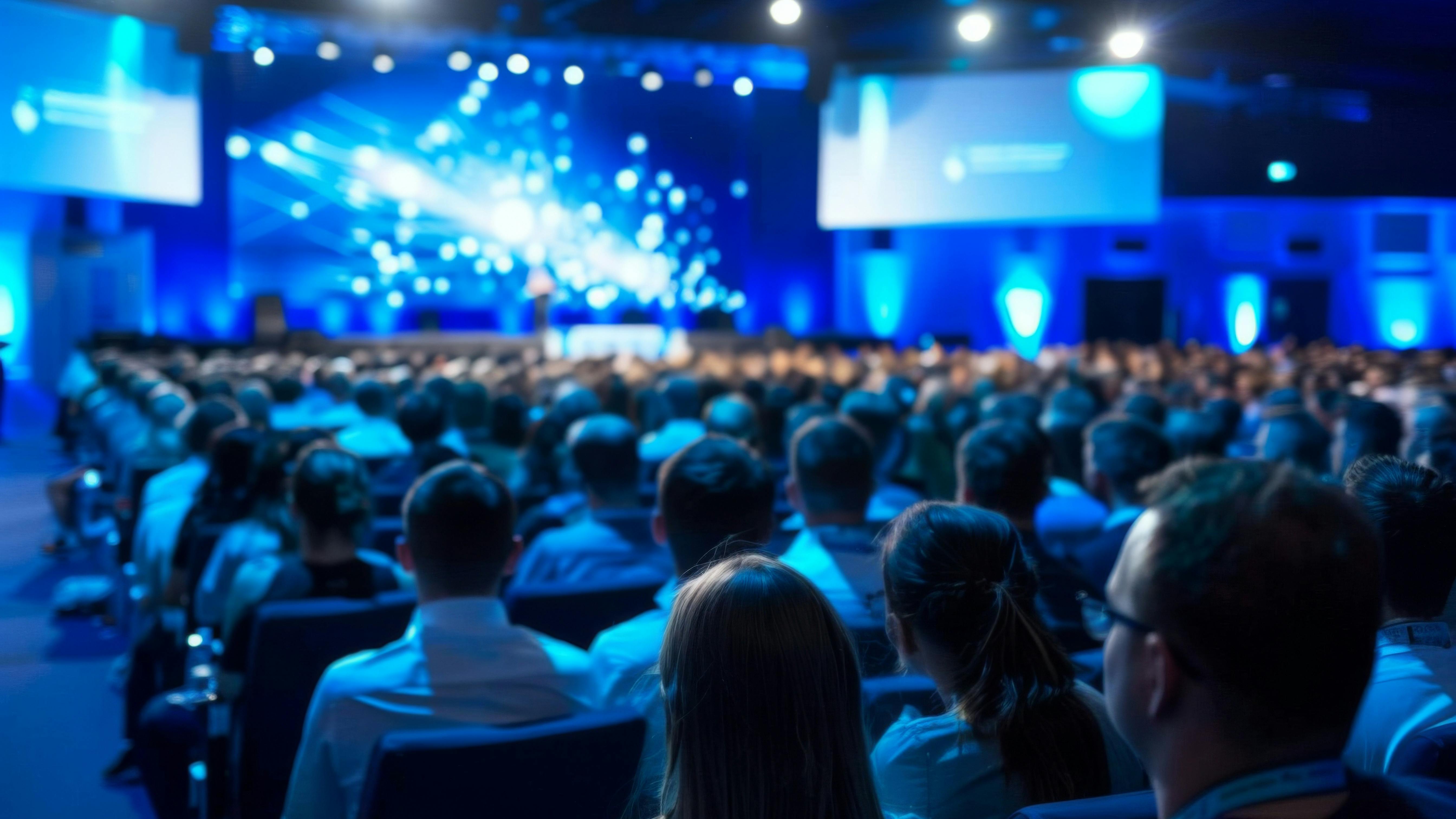 Blurred presenter addressing a large audience at a corporate conference event.