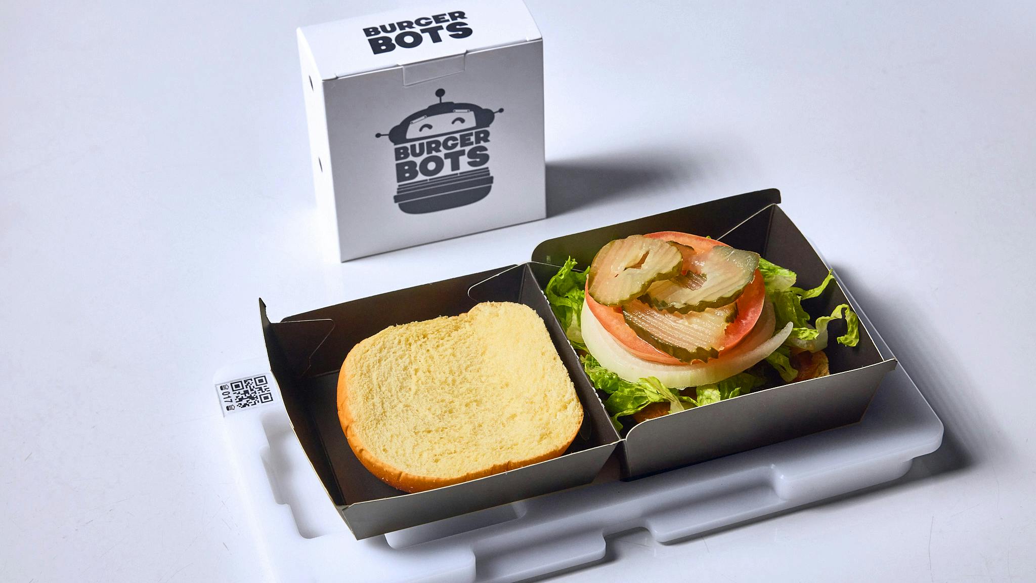 A BurgerBots burger box standing upright with another burger box open, showing an assembled burger inside, sitting on the QR code tray used in assembly on the shuttle conveyor.
